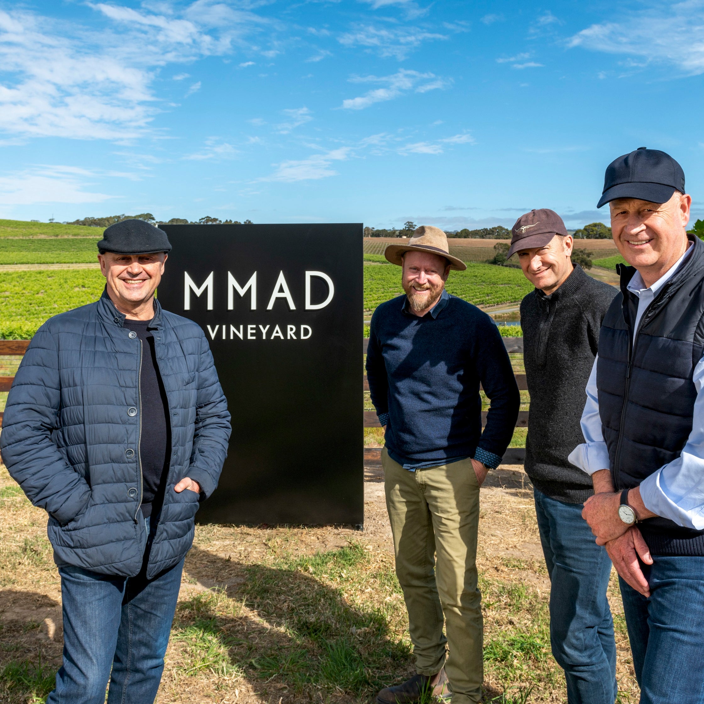 Four men standing in front of a sign for MMAD Vineyard with a vineyard in the background.